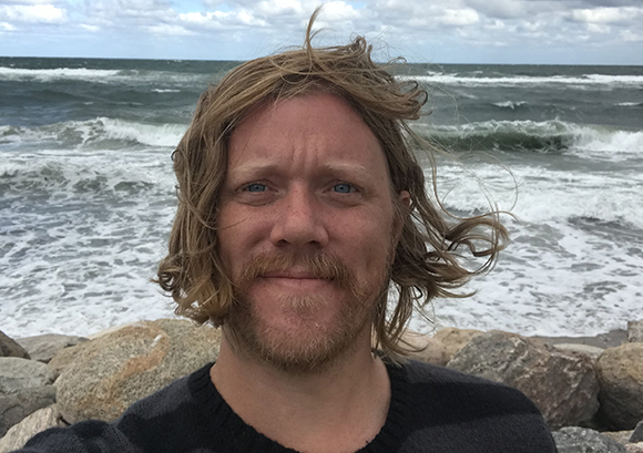 A man with wavy blond hair and a mustache looks at the camera with ocean waves in the background.