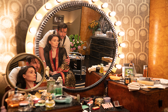A man leans toward a woman seated in front of a large, circular vanity mirror lined with bright light bulbs.