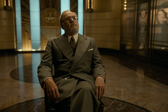 An older man with a grey beard and glasses sits in a grey double-breasted suit in a large, ornate hall.