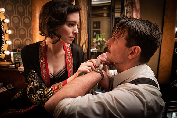 A woman in a black and red robe gazes intensely at a man in a white shirt and suspenders.