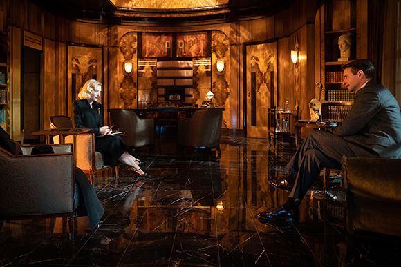 A woman in black sits across from a man in a suit in a dark, opulent wood-paneled room with marble floors.