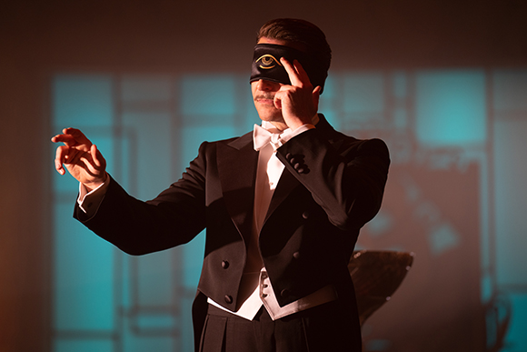 A man in a black tuxedo and white bowtie wears a black blindfold with a gold eye symbol.