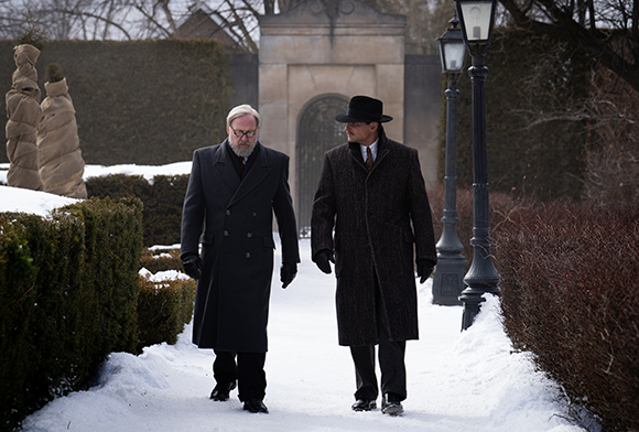 Two men in heavy overcoats and hats walk along a snow-covered path in a garden.