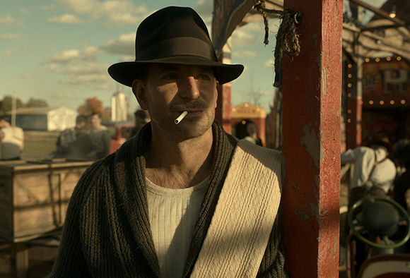 A man wearing a black fedora and a dark cardigan leans against a red post while smoking a cigarette.