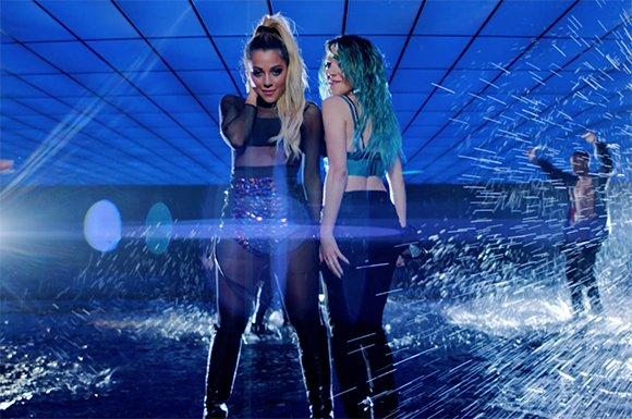 Two women in stylish outfits pose back-to-back as water splashes around them in a blue-lit setting.