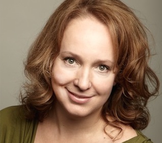 A woman with wavy reddish-brown hair and light eyes smiling against a neutral background.