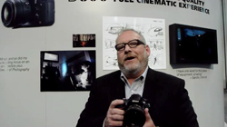 A man with glasses and a beard holds a black camera in front of a wall with photography displays.