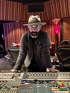 A bearded man wearing a grey hat and jacket leans over a large audio mixing console in a studio.