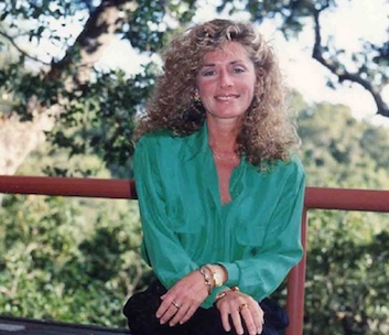 A smiling woman with long, curly blonde hair wearing a bright green long-sleeved blouse.
