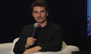 A man in a black blazer sits in a white chair holding a microphone.