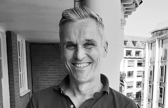 A smiling middle-aged man with short hair wearing a polo shirt outdoors.