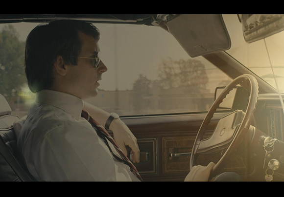 A man wearing sunglasses and a tie sits in the driver's seat of a vintage car.