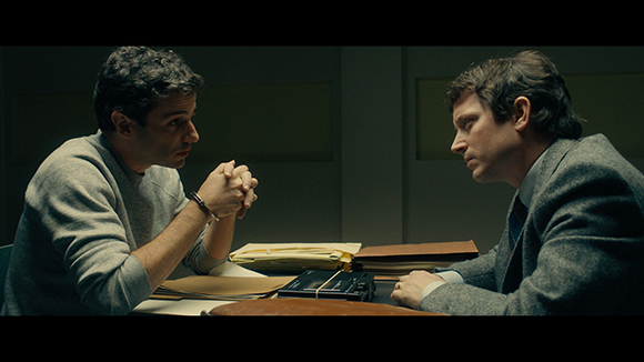 Two men in a dimly lit room sit across a table with files and a tape recorder between them.