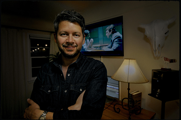 A man with dark hair and a beard wearing a denim shirt, smiling in a dimly lit room.