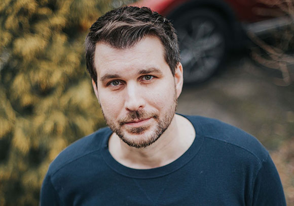 A man with short brown hair and a light beard wearing a dark blue crewneck shirt.