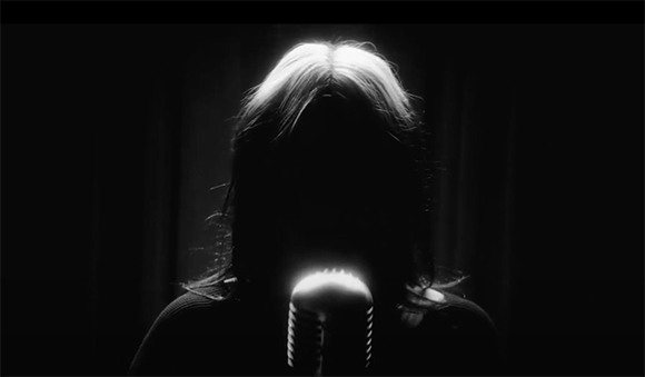 Silhouette of a person with long hair singing into a vintage-style microphone against a dark background.