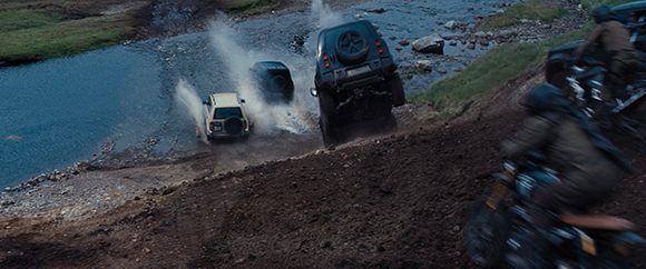 Off-road vehicles splash through a shallow river while motorcyclists ride along a dirt embankment.