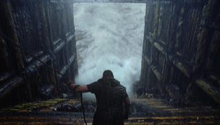 A man stands in a dark wooden structure looking out at large, white crashing waves.