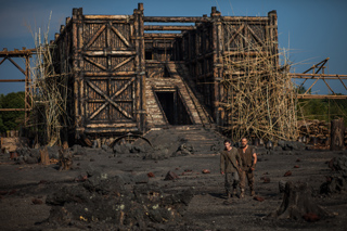 A large, weathered wooden structure sits in a dark, rocky field with two people standing nearby.