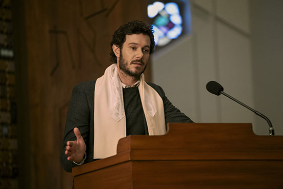 A man with dark curly hair and a beard wearing a light pink stole stands behind a wooden podium.