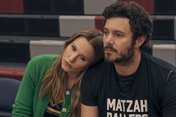 A woman in a green cardigan rests her head on the shoulder of a bearded man in a black t-shirt.