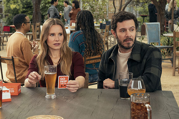 A man and woman sit at a wooden table outdoors, looking toward the side with surprised expressions.