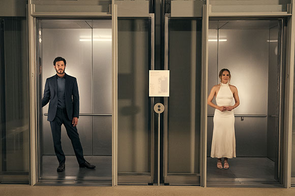 A man in a blue suit and a woman in a white dress stand inside two adjacent open elevator cars.