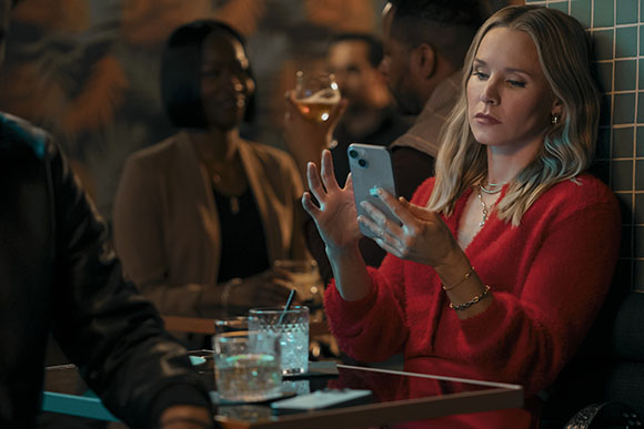 A blonde woman in a red sweater looks at her smartphone while sitting at a bar with people in the background.