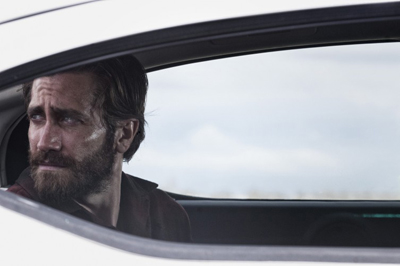 A bearded man looks out of a white car window with a serious expression.