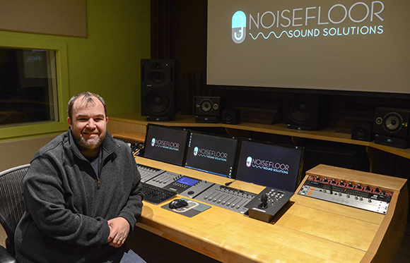 A man sits in front of a mixing console and multiple monitors in a sound studio.