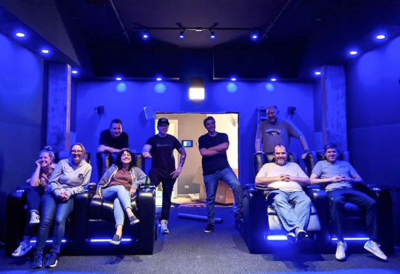 A group of ten people smiling and posing in a dark room with blue ambient lighting and cinema seating.