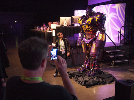 A person holds up a smartphone to take a picture of a large, purple and gold robot with glowing lights. Another person stands