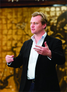A man in a black blazer and white shirt gestures with his hands while speaking in front of a gold background.