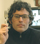 A man with curly hair and glasses holds a silver fork up near his face.