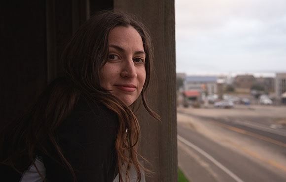A woman with long brown hair looks toward the camera from behind a dark wooden structure.