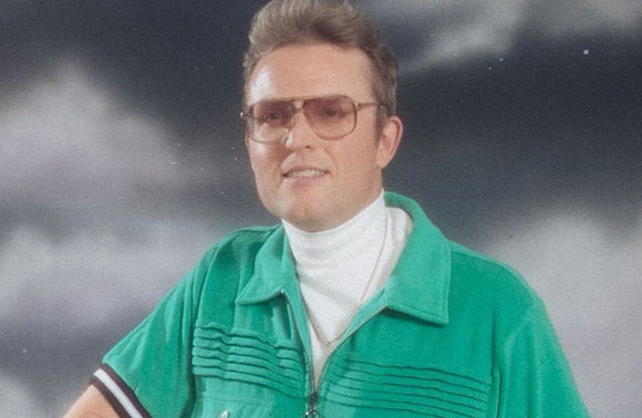 A man with light brown hair wearing gold-rimmed glasses, a white turtleneck, and a green zip-up shirt.