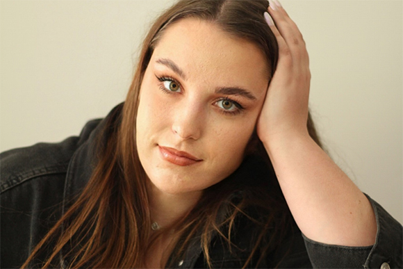 A young woman with long brown hair looking at the camera with her hand resting on her head.