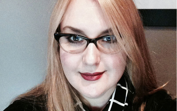 A woman with long blonde hair and black-rimmed glasses smiling slightly.