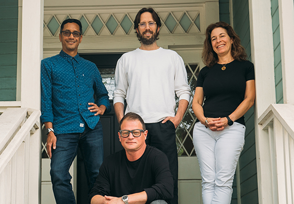 Four people standing and sitting on a white porch in front of a house.