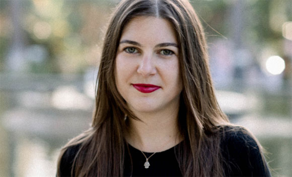 A woman with long brown hair and red lipstick looks toward the camera against a blurred outdoor background.
