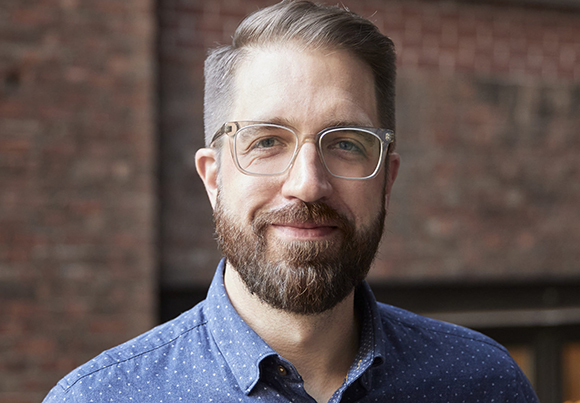 A man with short brown hair, glasses, and a brown beard smiling in front of a brick wall.