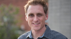 A man with short brown hair smiling at the camera in front of a blurred outdoor background.