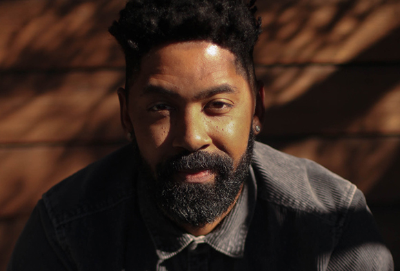 A man with dark curly hair and a salt-and-pepper beard looks toward the camera against a wooden wall.