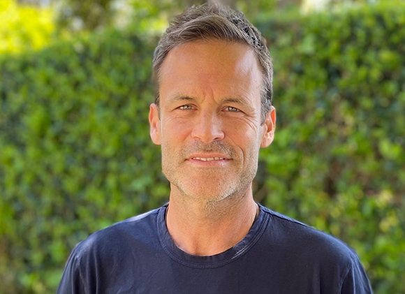 A man with short brown hair and a slight smile wearing a dark t-shirt against a blurred green background.