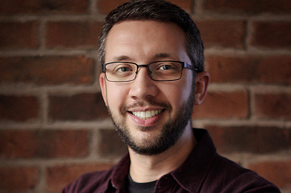 A man with short dark hair, glasses, and a beard smiling against a red brick background.