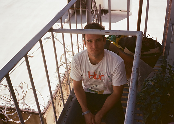 A man in a white t-shirt sits on outdoor stairs next to a metal railing and potted plants.
