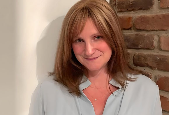 A woman with shoulder-length reddish-brown hair and bangs smiling against a white wall and brick background.