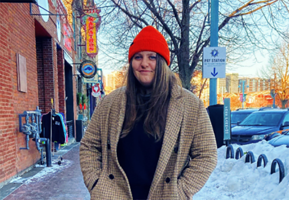 A woman wearing a red beanie and a tan plaid coat stands on a snowy city sidewalk near a brick building.