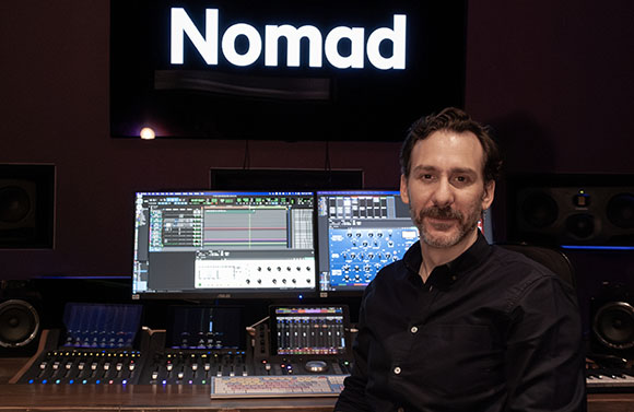 A man sits in a recording studio in front of computer monitors and a mixing console.