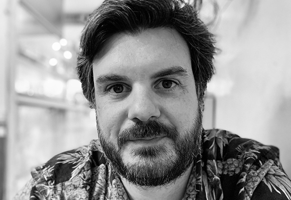 Black and white close-up portrait of a man with dark hair and a beard wearing a patterned shirt.
